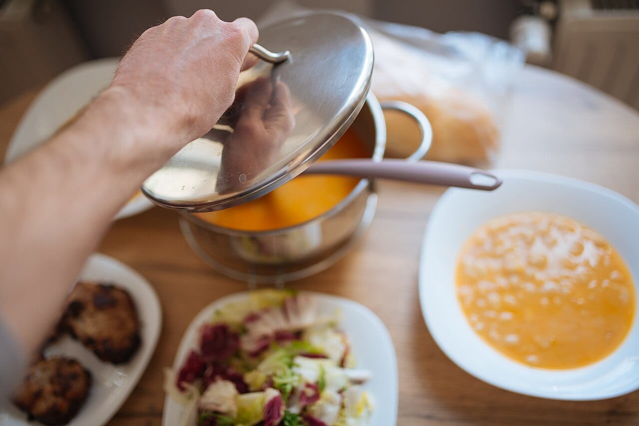 A person stirs a pot of soup on a kitchen counter with salad and bread nearby