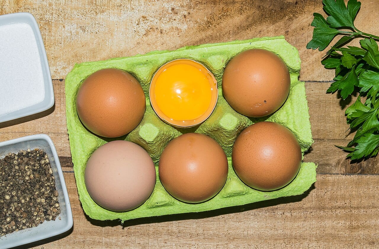 A green carton of six brown chicken eggs, one of them opened to its yolk