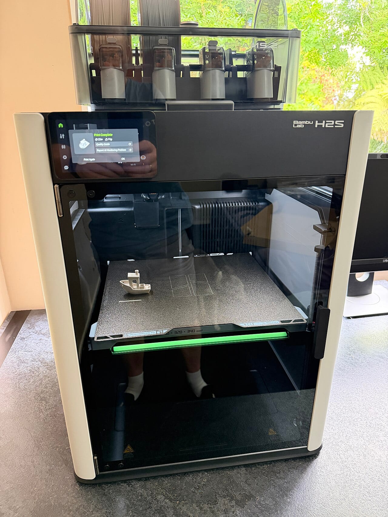 A Bambu Lab H2S 3D printer on a desk, its print bed visible through the front door