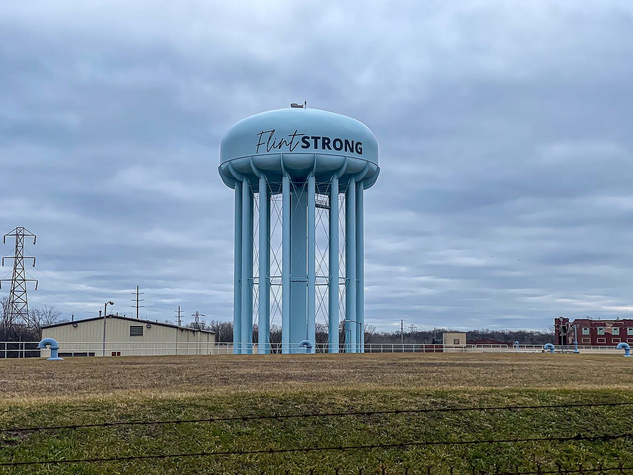 A blue water tower painted Flint Strong stands over a field in Flint, Michigan, under a gray sky