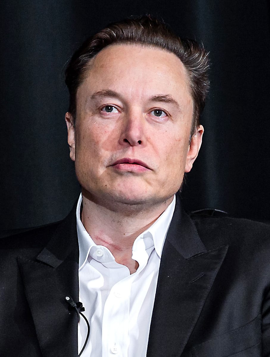 Elon Musk in a dark jacket and white shirt, looking at the camera with a serious face