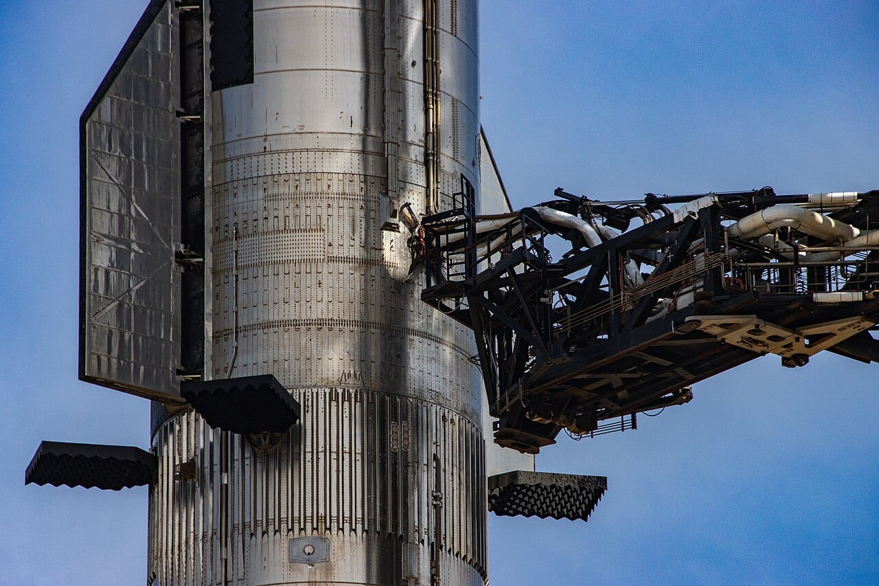 A Starship stacked on its Super Heavy booster, with the launch tower's quick-disconnect arm reaching toward it against a blue sky