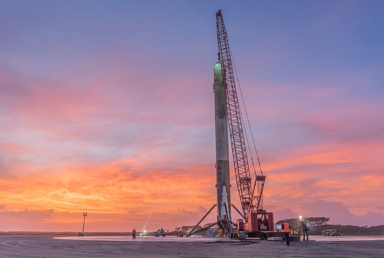 A Falcon 9 first stage standing upright on Landing Zone 1 beside a crane at sunset