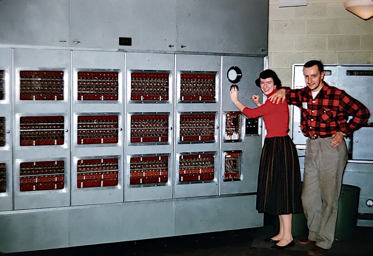 A smiling woman in a red sweater and a man in a plaid shirt stand beside the gray metal cabinets of the ILLIAC I computer