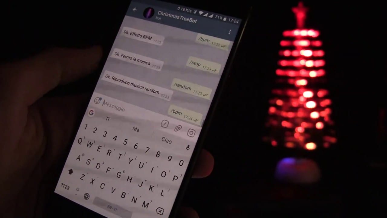 A hand holds a phone open to a Telegram bot chat, beside a lit Christmas tree the bot controls